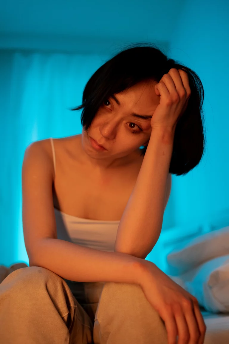Femme asiatique assise sur le bord du lit, de face, avec un fond bleu, la tête posée sur sa main l'air épuisée et déprimée