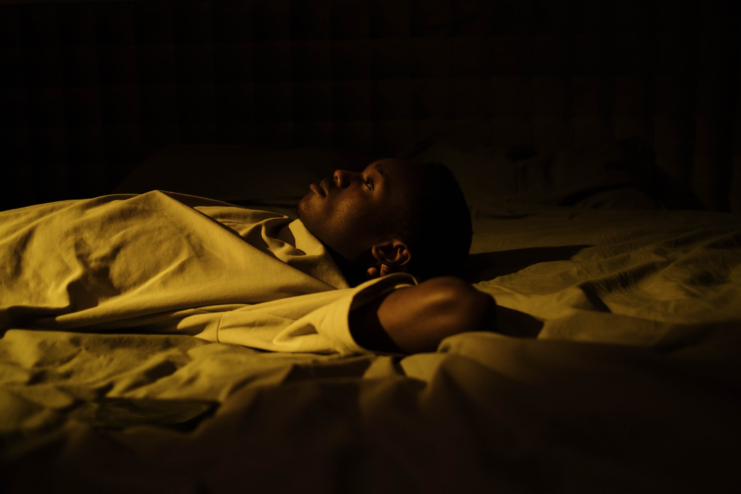 Un homme noir allongé sur un lit, les bras croisés derrière la tête, réveille, habillé en blanc dans une lumière jaune