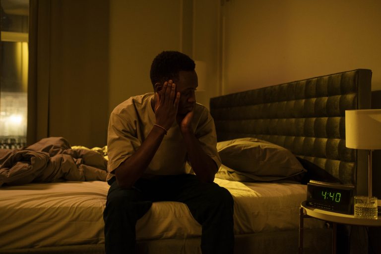 Homme noir assis sur le bord du lit, avec un t-shirt et un jean, en train de regarder son réveil qui indique 4 heures du matin