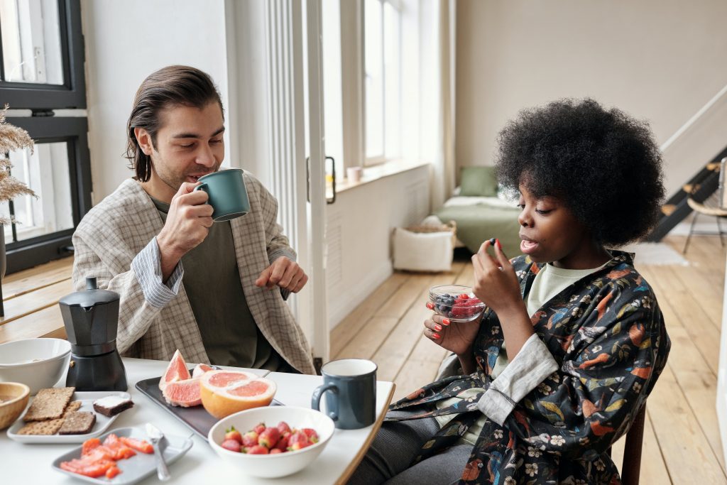 Un homme blanc et une femme noire prennent un café en riant, devant un petit-déjeuner de fruits